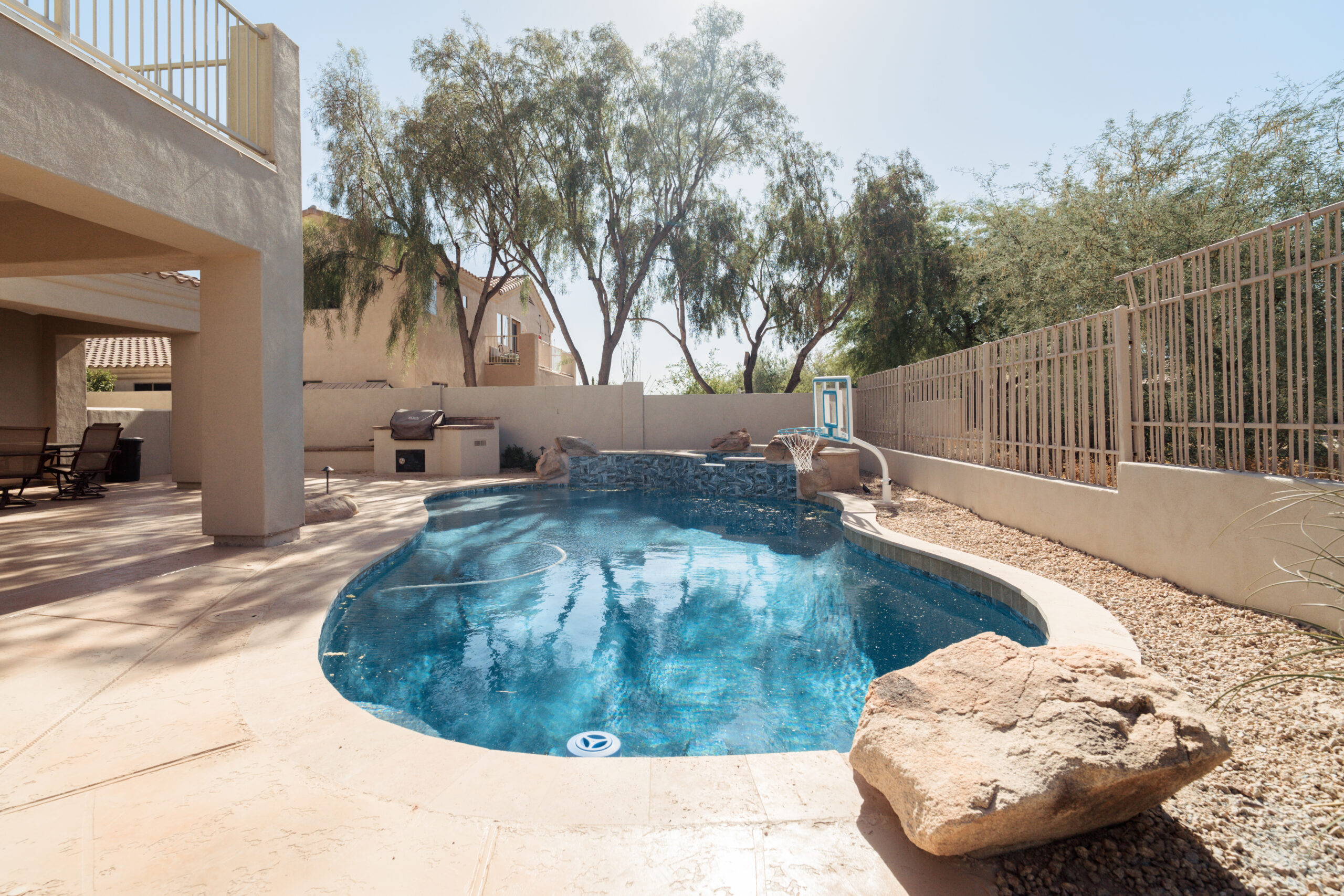 Curved outdoor swimming pool with a stone patio, nearby outdoor grill, and a beige fence.