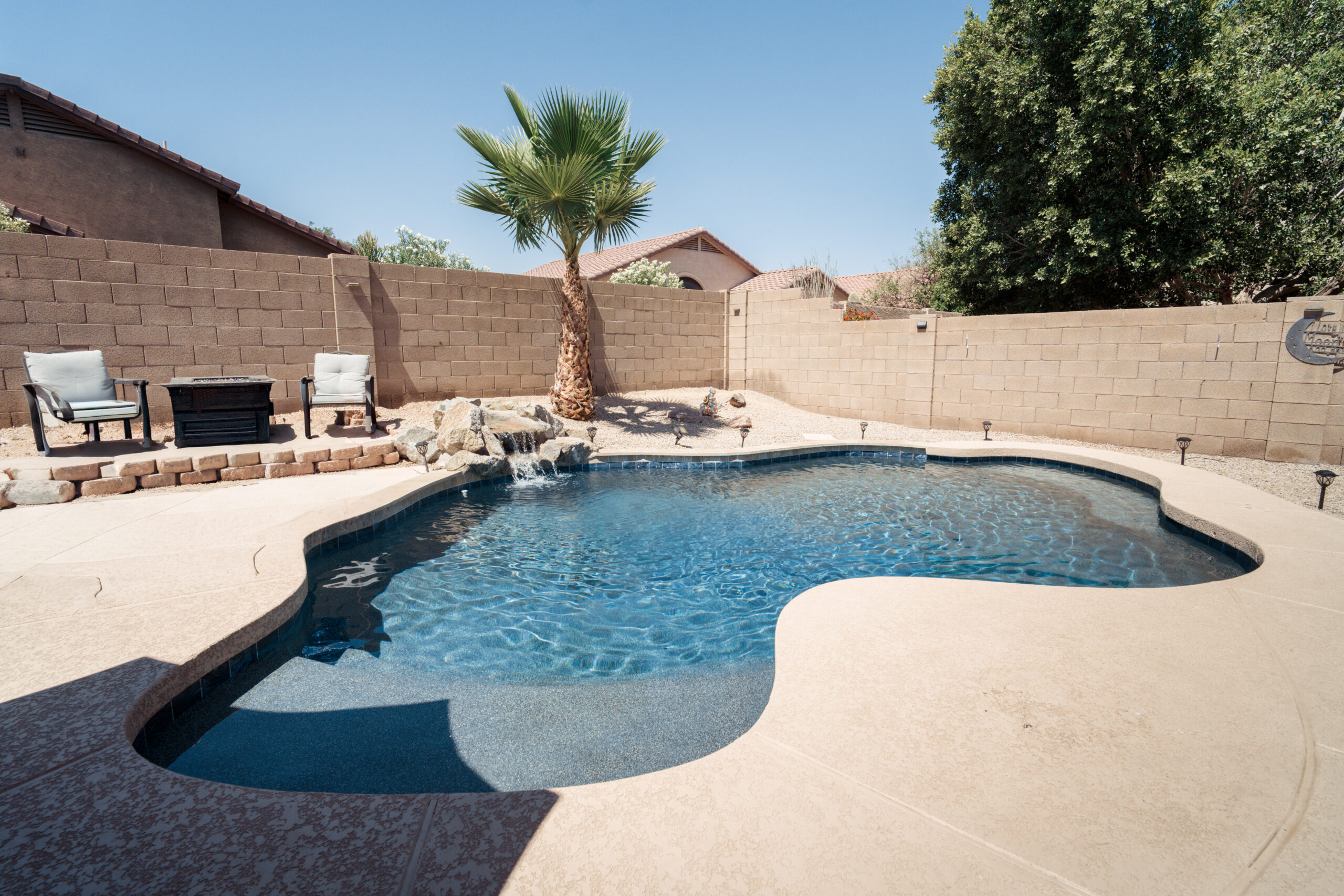 Freeform backyard swimming pool with a rock waterfall, patio seating, and a palm tree against a tan block wall.