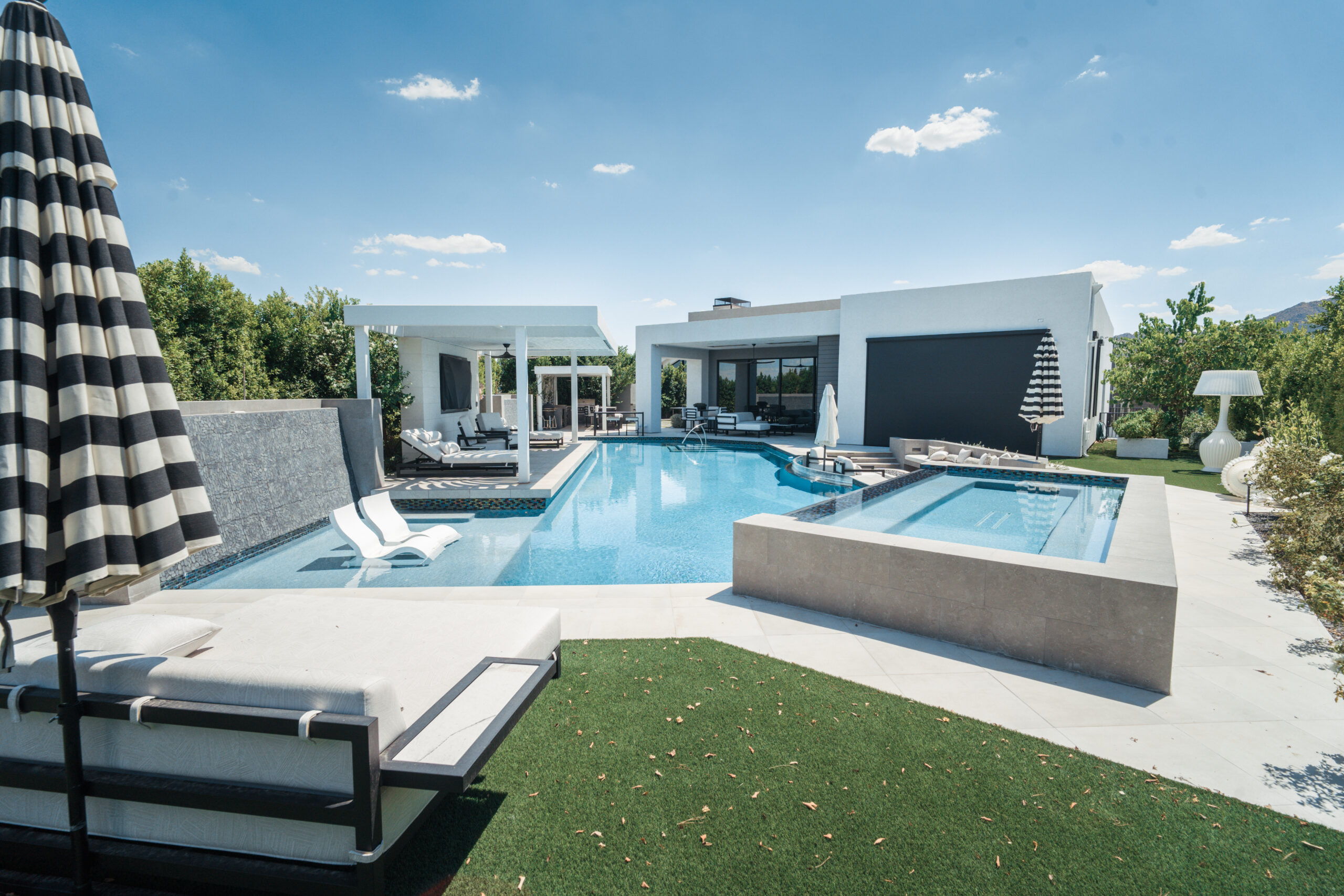 Modern outdoor patio with a swimming pool, integrated spa, lounge chairs, and a minimalist white house under a blue sky.