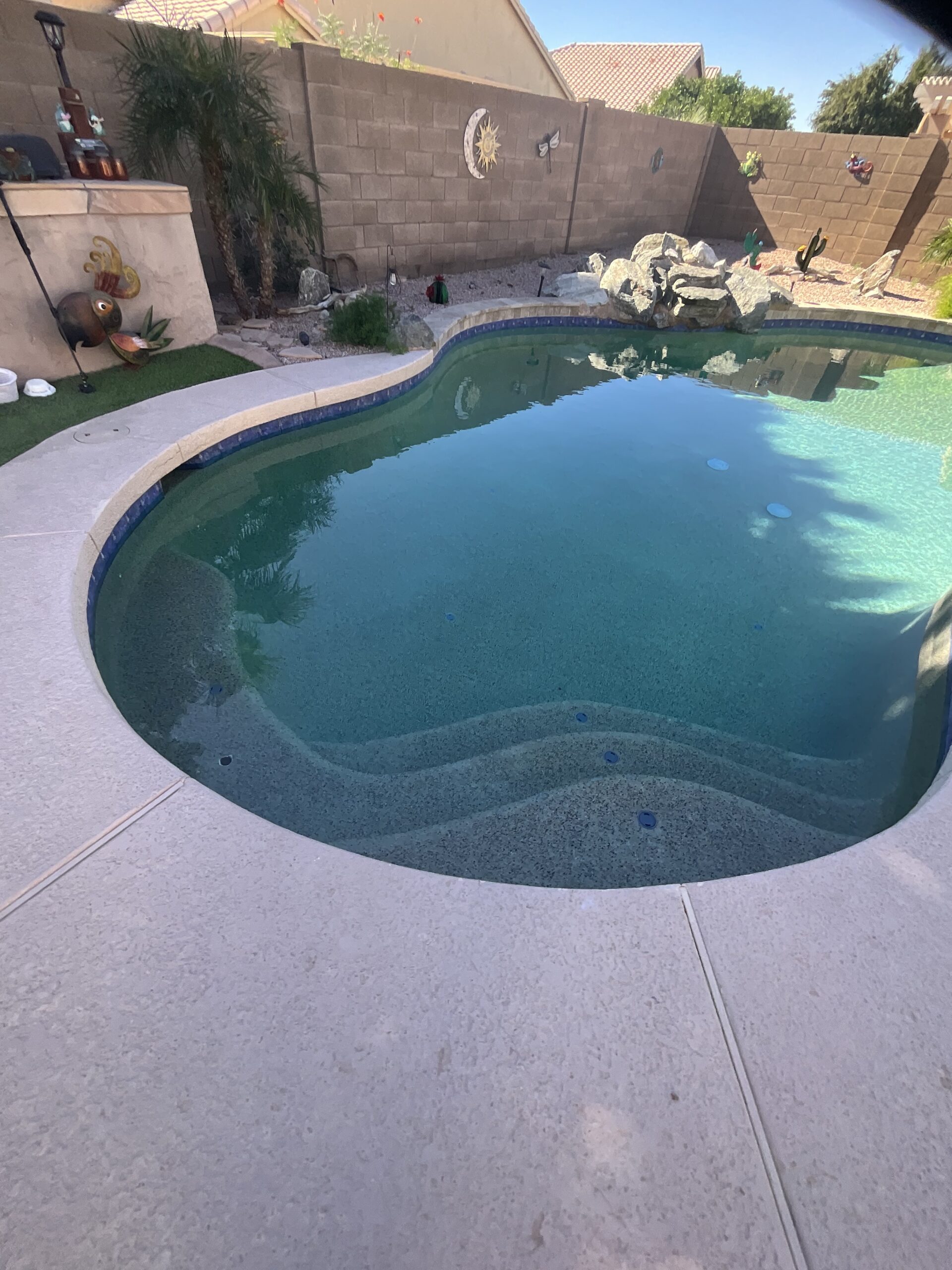 Curved swimming pool with built-in steps, surrounded by a concrete patio, a rock garden, and a tan stone wall.