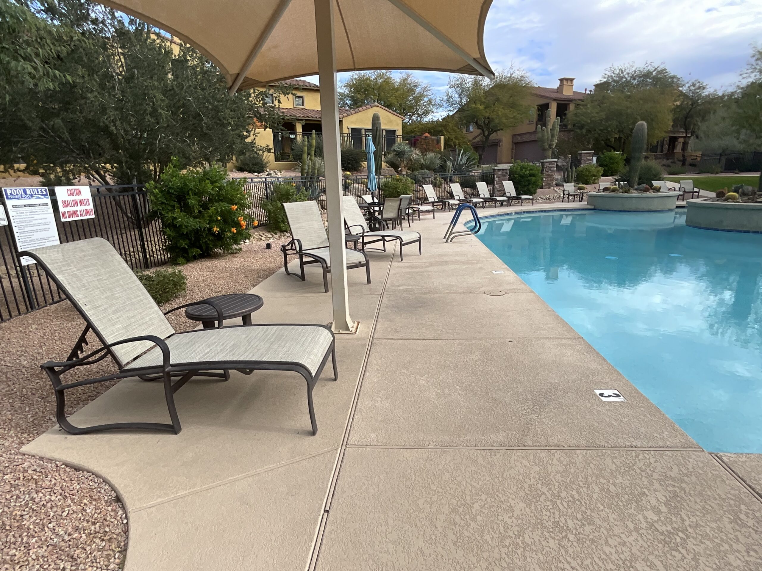 Outdoor swimming pool area with lounge chairs under a large beige umbrella and a pool rules sign.