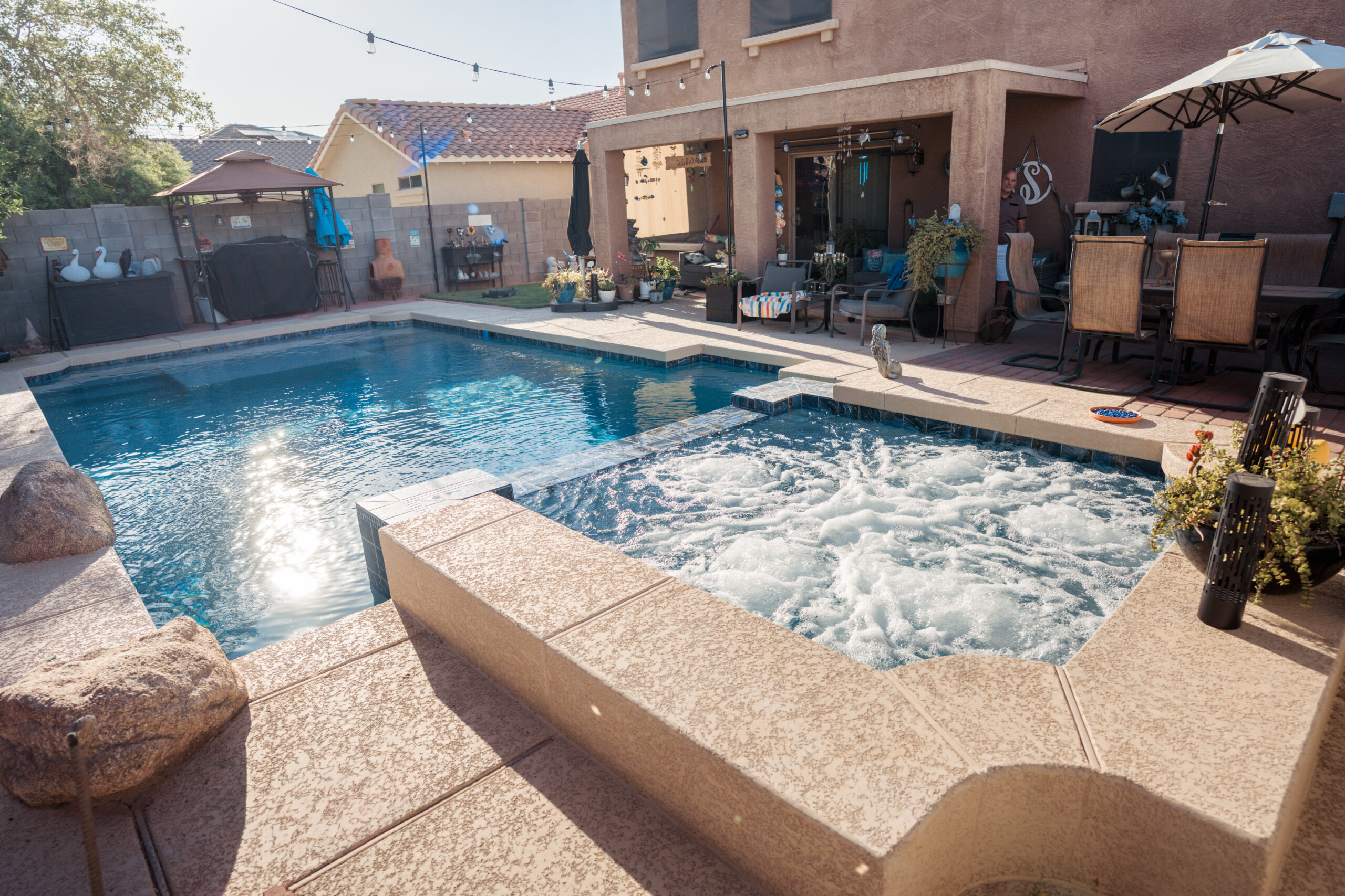 Backyard patio with a swimming pool, bubbling hot tub, outdoor dining area, and a tan stucco house.
