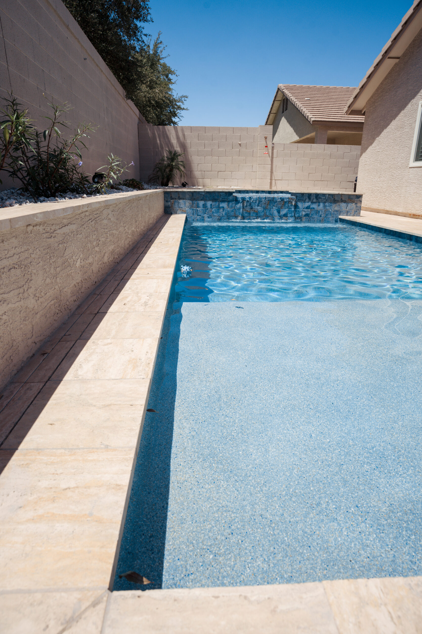 Narrow lap pool with light blue tiling, tan stone decking, and a small waterfall feature in a walled backyard.